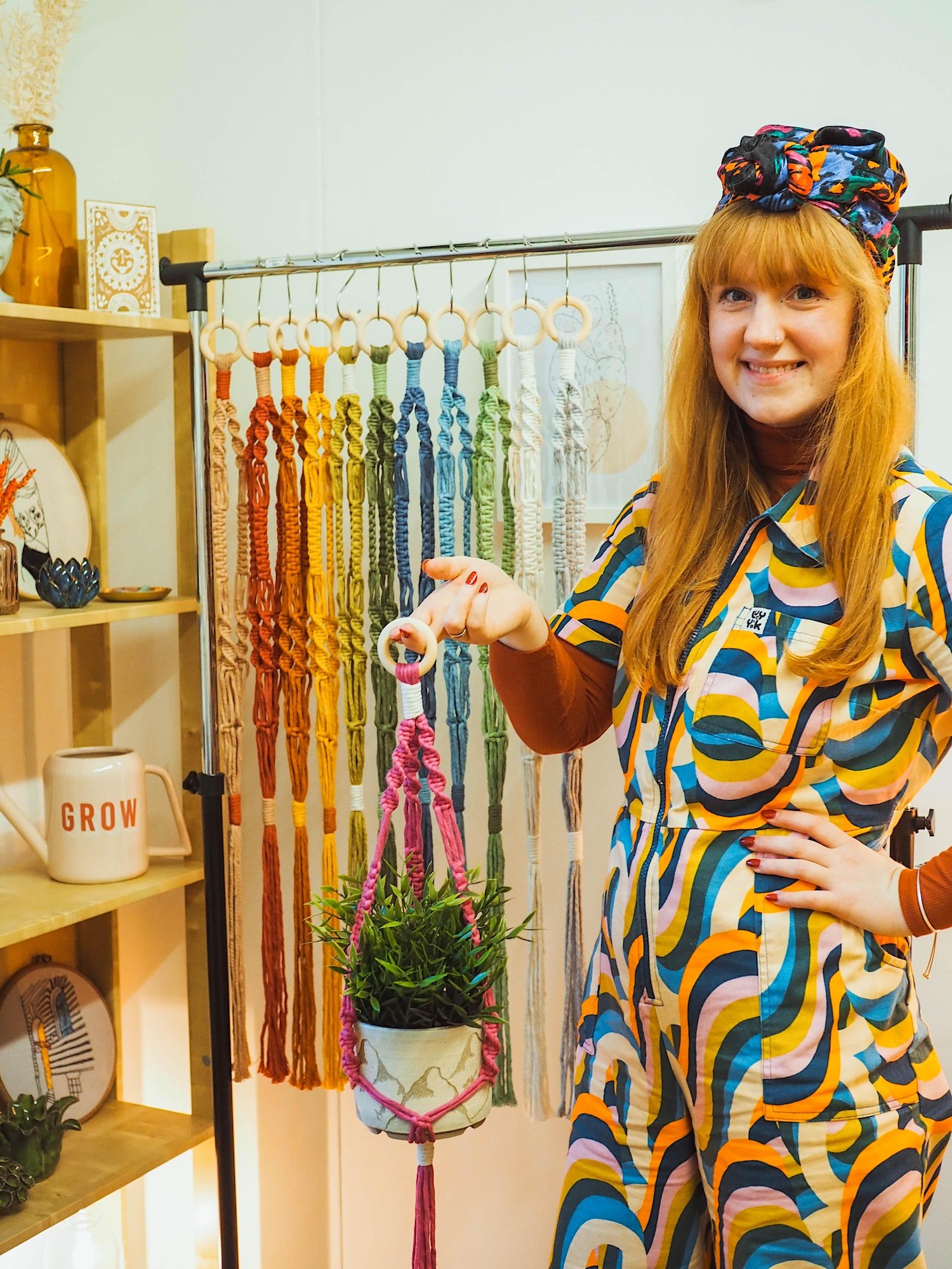 A ginger haired woman in a geometrical printed jumpsuit holds a plant in a pink macramé hanger