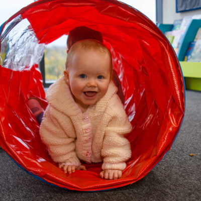 A smiling white baby wearing a pink jumper crawls through a res play tunnel looking at the camera