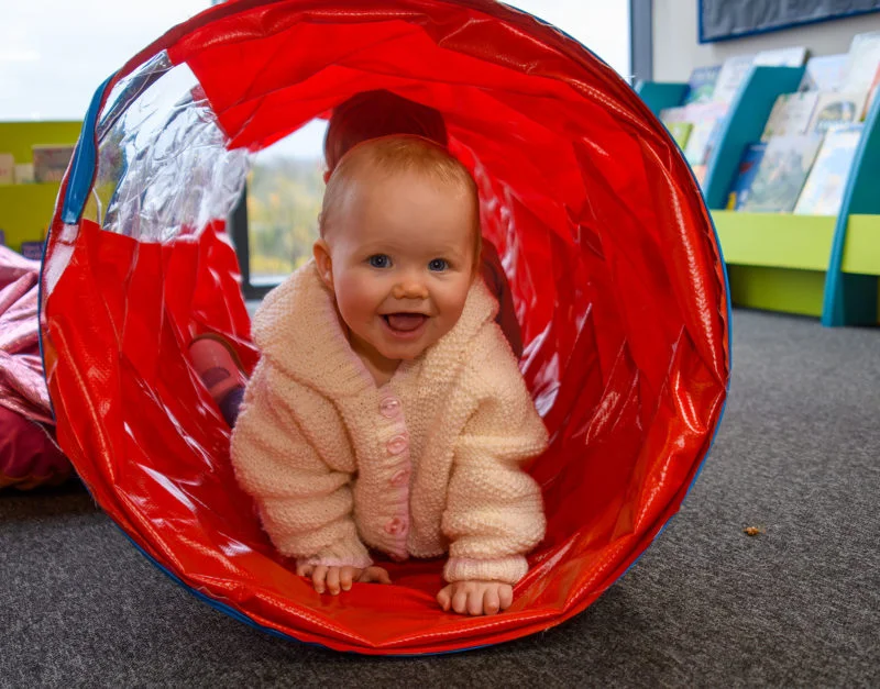A smiling white baby wearing a pink jumper crawls through a res play tunnel looking at the camera