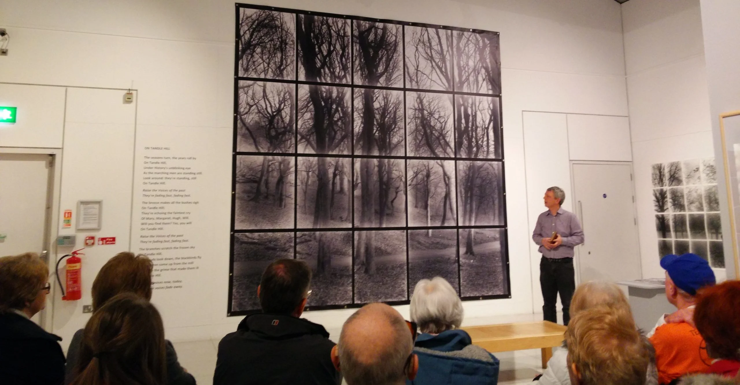 A tall paneled photographic exhibition with a man stood to the side giving a talk