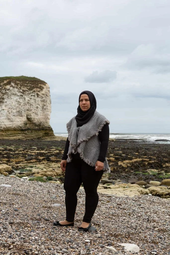 A woman wearing a black headscarf stands on a pebbled beach with a white cliff edge in the background