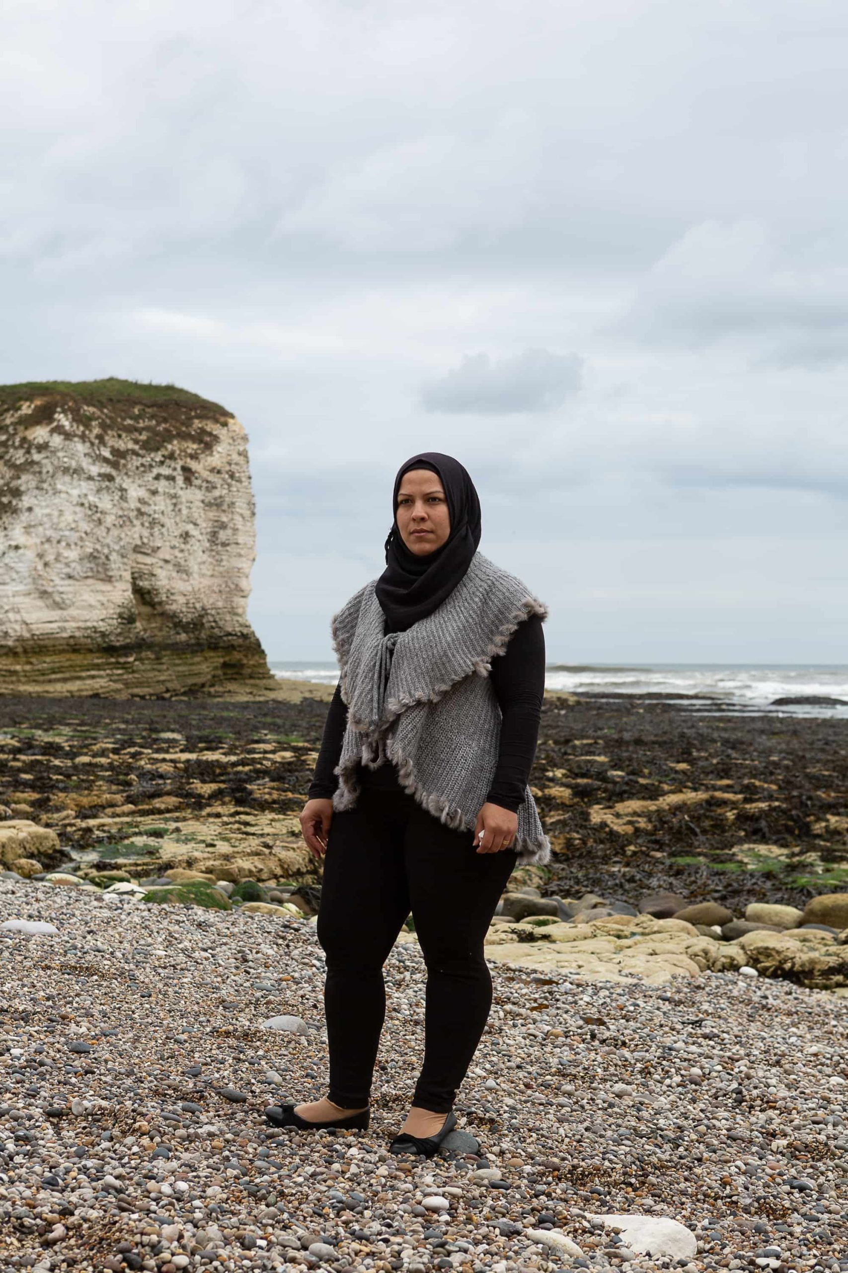 A woman wearing a black headscarf stands on a pebbled beach.