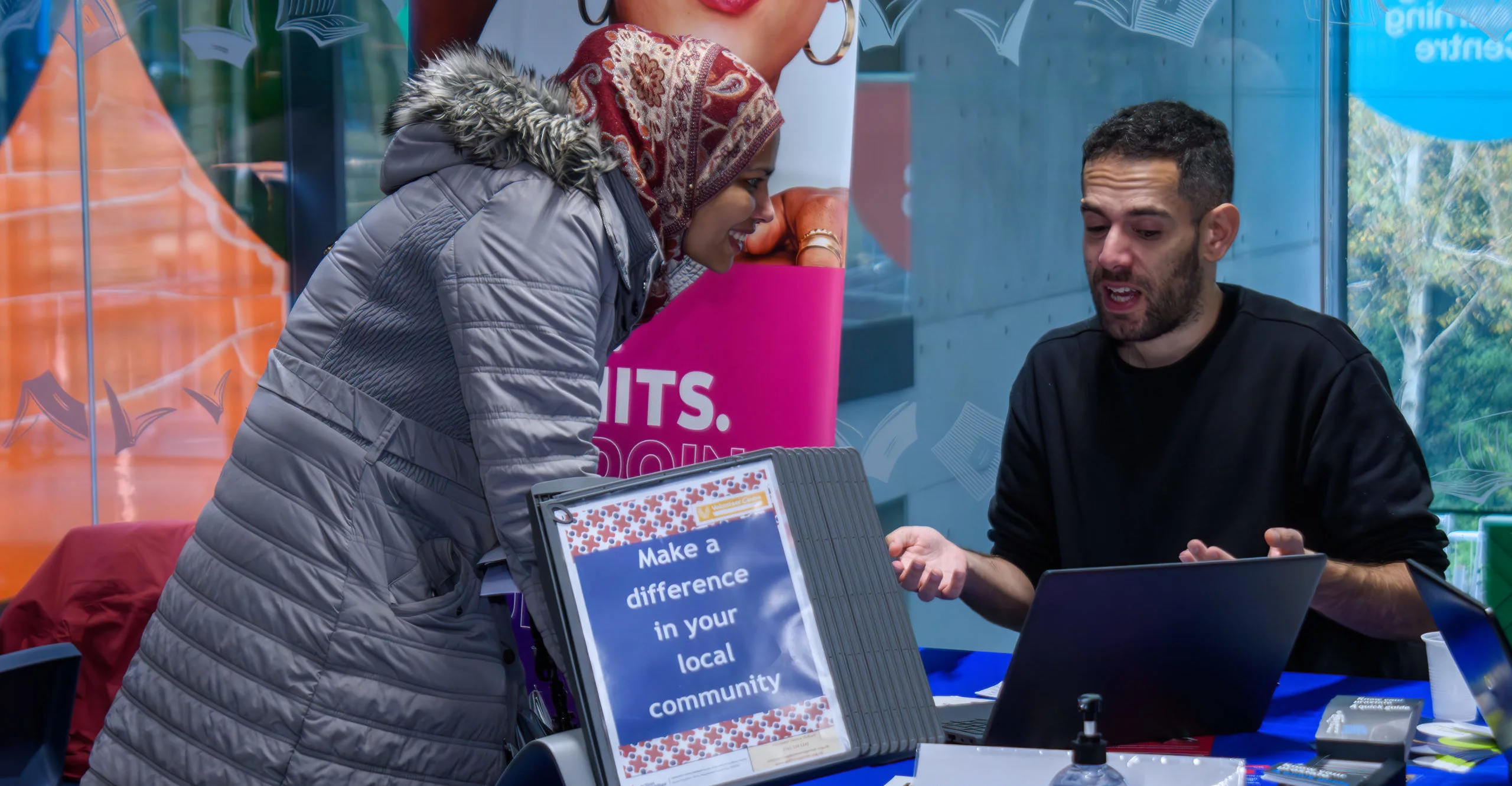 A woman wearing a headscarf leans in smiling towards a laptop next to a man in a black jumper