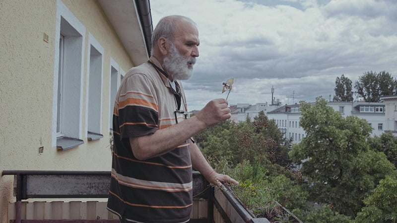 Bani Abidi, The Song, 2022, film still showing a man on a balcony blowing a mini paper windmill.