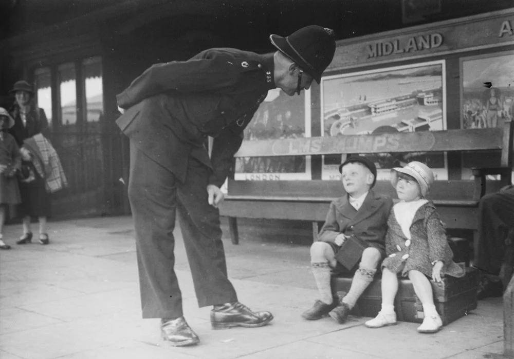Police officer looking down at a boy and girl sat on a suitcase at the train station about 1930s.