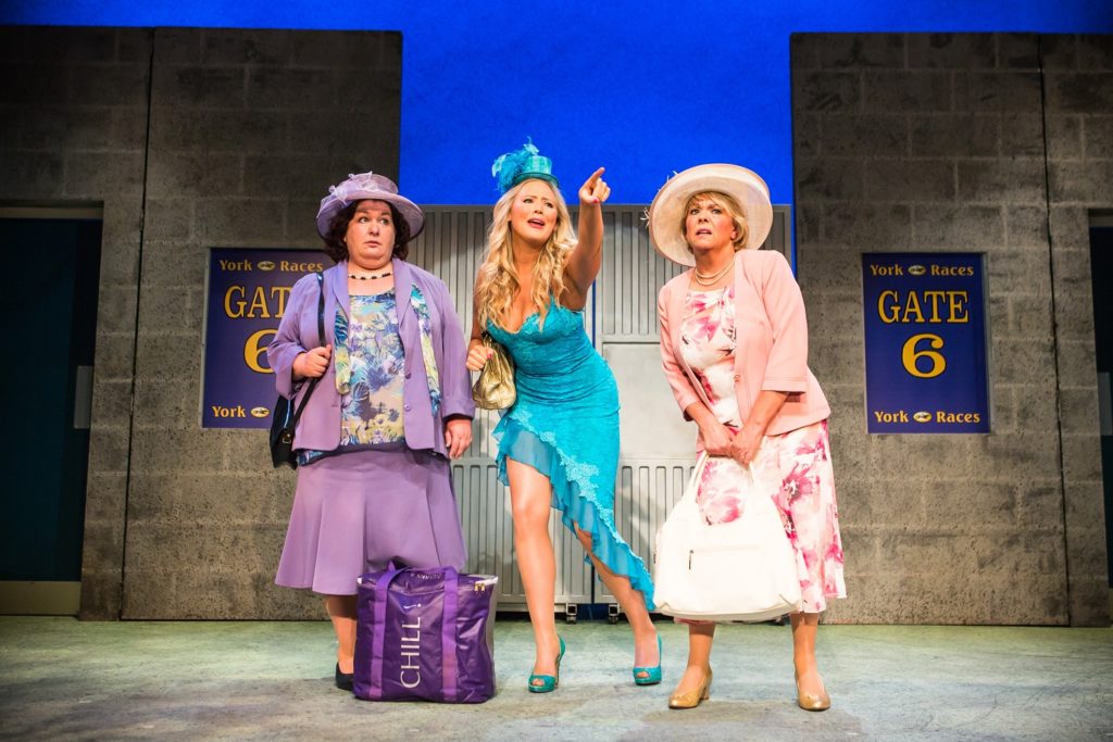 Three women stood on stage with a backdrop of the gates at York Races. They are dressed up and in hats.