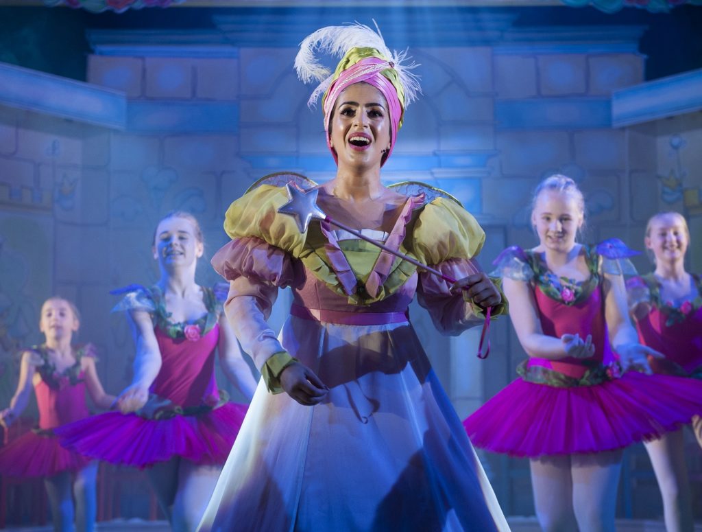 Fairy godmother singing and holding wand with four girls dancing behind.